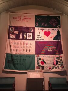 A memorial quilt hanging vertically on a wall, made up of patches running two wide and four long. Some are minimally decorated, like a tombstone, while others are adorned with photo collages. One patch memorializes 21 lives alone, each represented by a candle labeled with initials and birth and death dates.