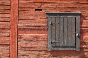 A closeup on a burnt orange, wooden wall. A window, framed in grey wood, is closed and padlocked.