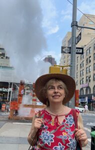 Picture of Sofi Stambo: A blond woman wearing a crimson, floral-patterned shirt and a wide-brim hat smiles, her eyes gazing up-and-right. She stands in front of a city intersection under construction, steam rising from the site.