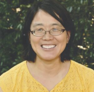 Haitao Xu's headshot: A woman with a black bob and oval-shaped glasses beams at the camera. She is wearing a yellow shirt and stands in front of dense green foliage.