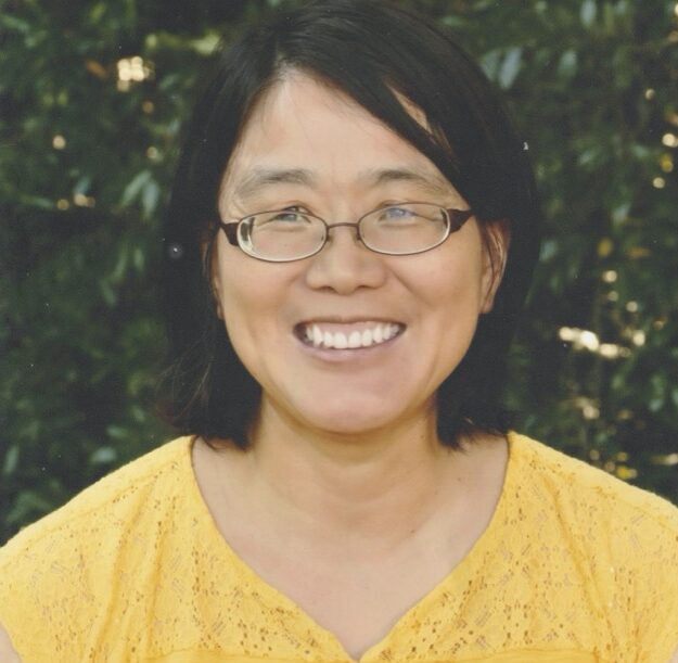 Haitao Xu's headshot: A woman with a black bob and oval-shaped glasses beams at the camera. She is wearing a yellow shirt and stands in front of dense green foliage.