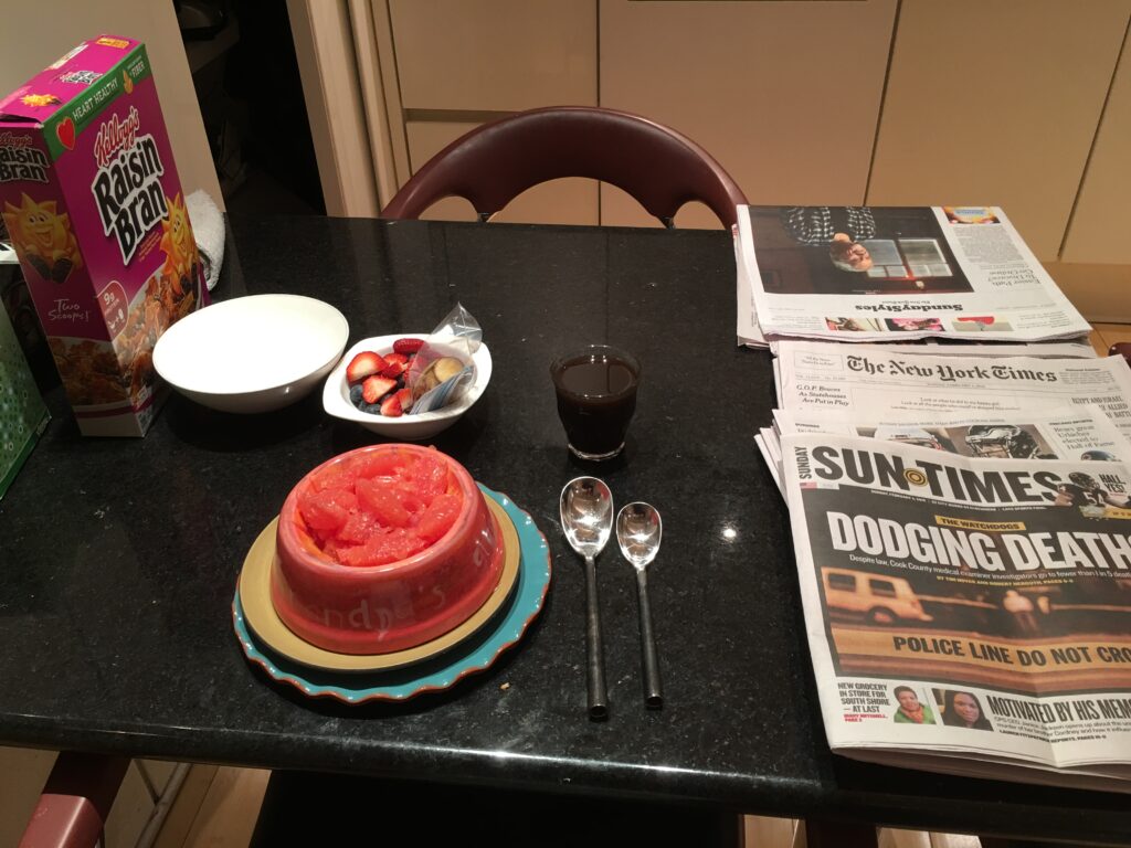 A small table set for breakfast: mashed grapefruit, berries, a Raisin Bran box, two spoons, and a short glass of dark liquid. To the right of the place setting is a stack of newspapers, including the Chicago Sun Times.