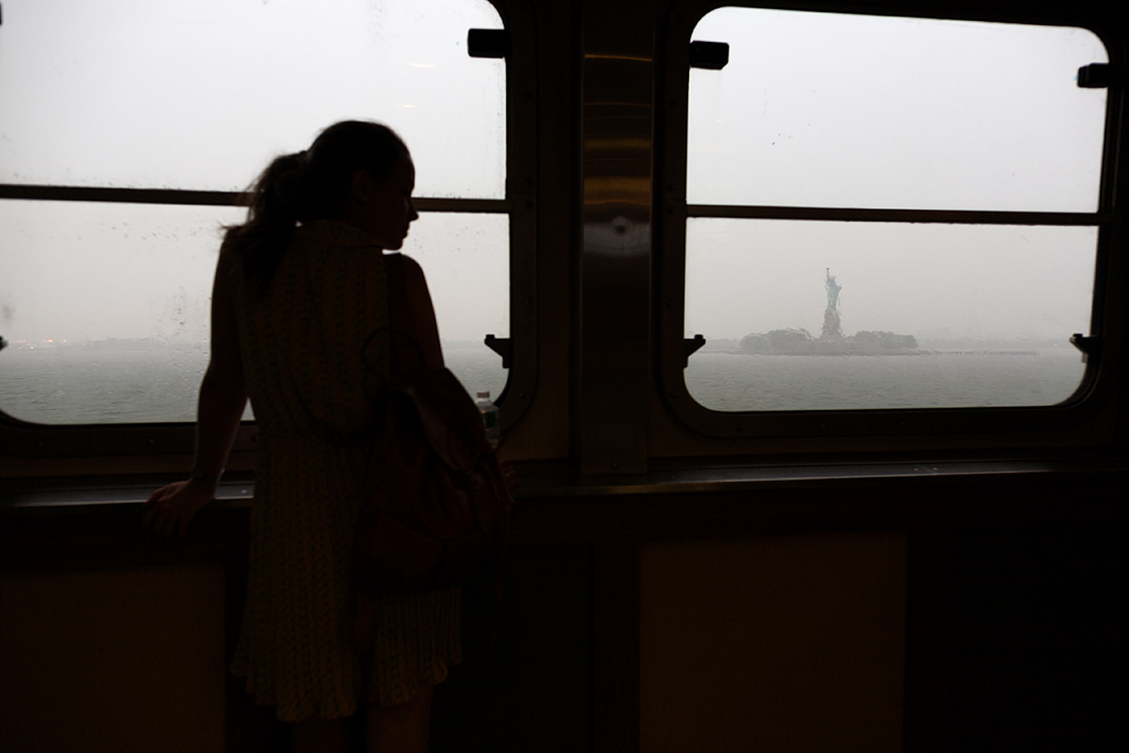 A woman stands in front of a train window, the statue of Liberty visible in the distance.